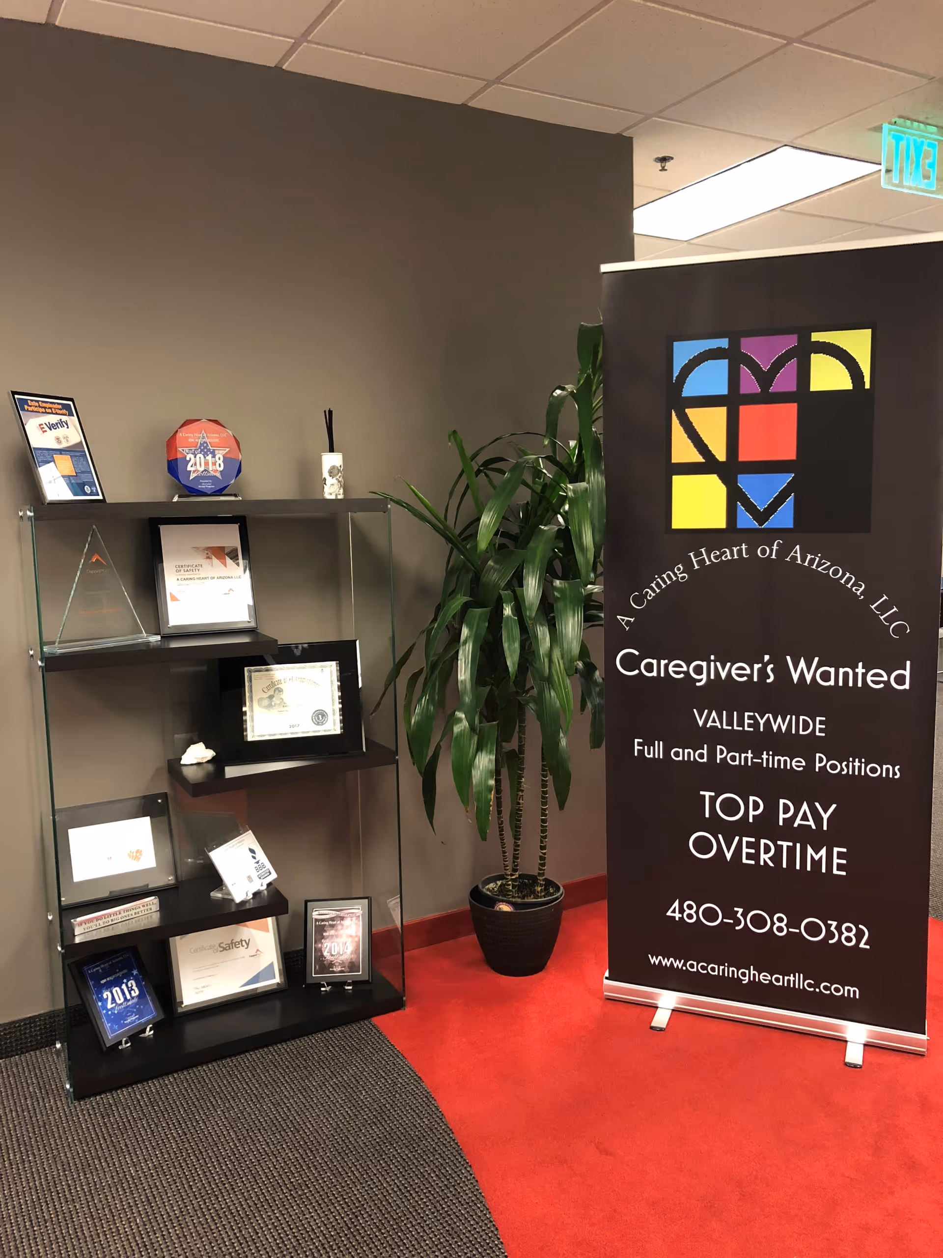 Indoor lobby corner featuring a tall banner, a potted plant, and a display shelf with awards.