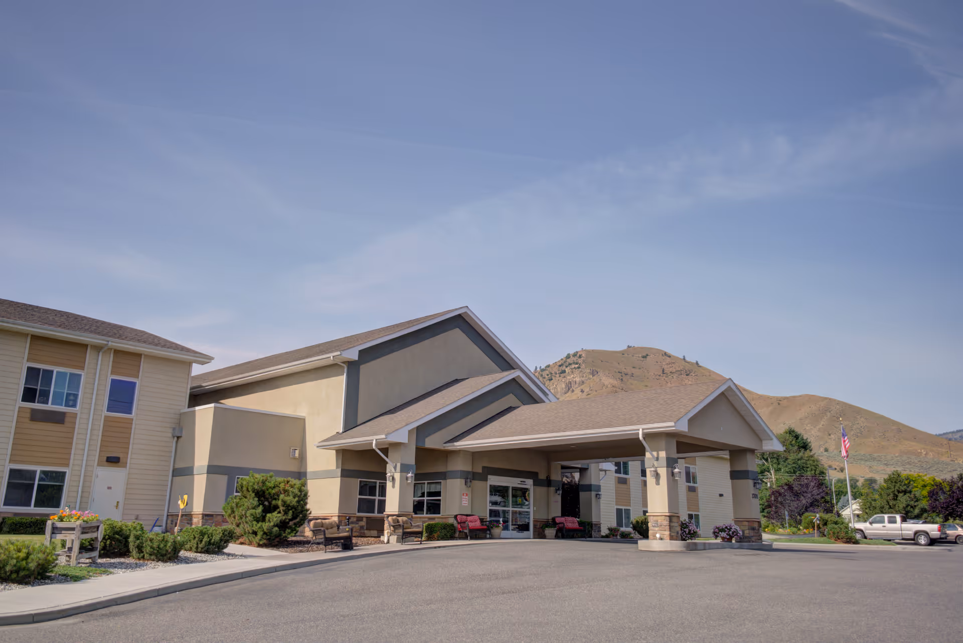 Exterior view of Blossom Valley by Cogir senior living facility with a covered entrance, benches, and a parking area. The building is set against a backdrop of hills under a clear blue sky.
