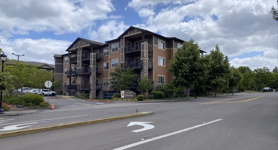 Three-story senior living building, The Springs at Tanasbourne, with balconies, trees, and a street in front.