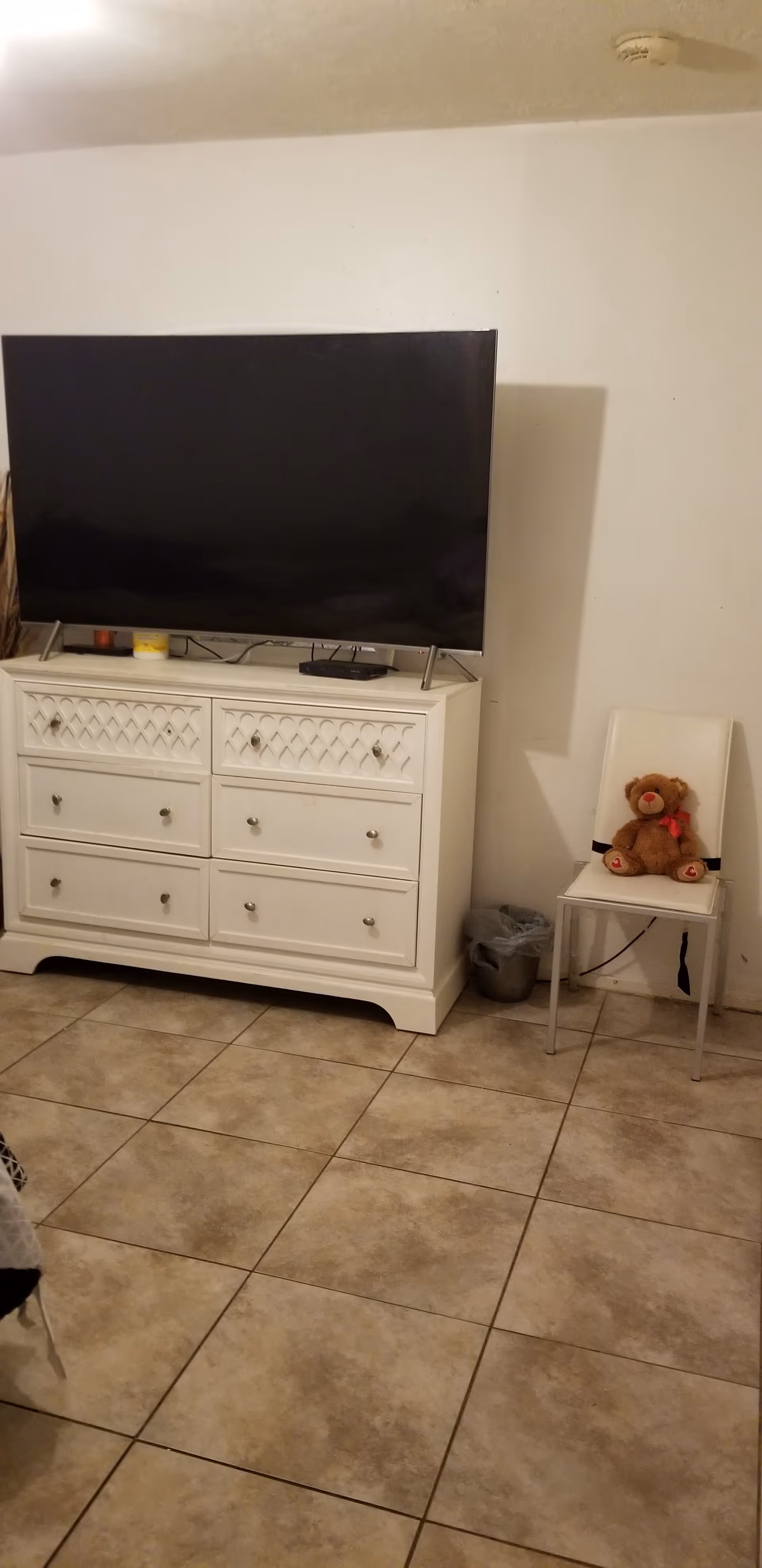 White dresser with flat-screen TV, small chair with teddy bear, tiled floor.