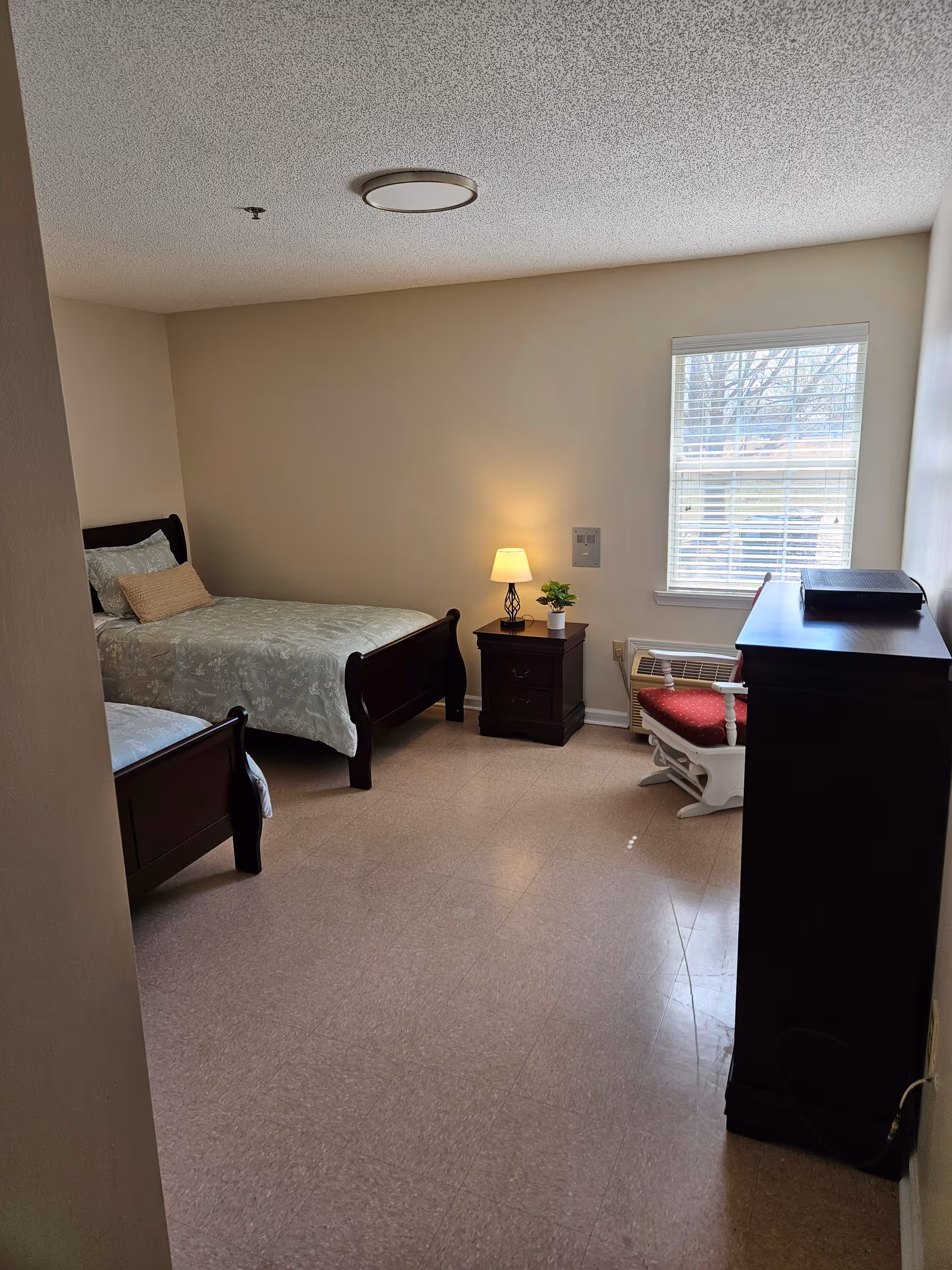 Two twin beds, a nightstand with lamp, dresser, and windowed corner in a senior living bedroom.
