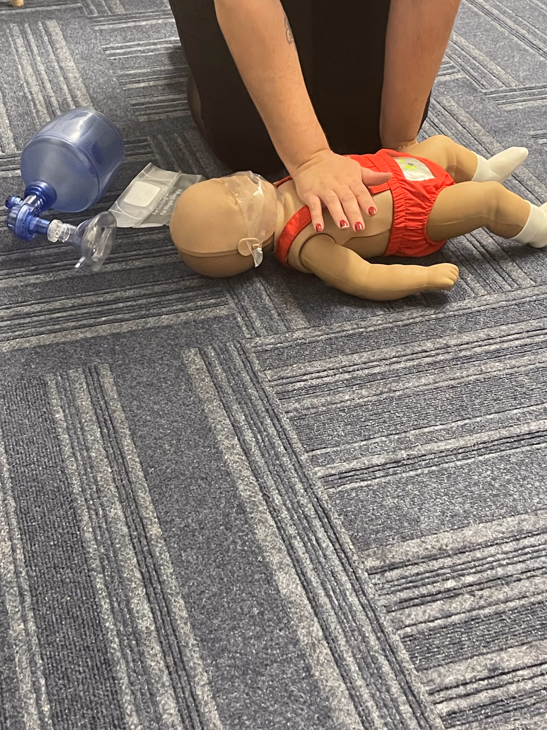 A CPR training mannequin lies on carpeted floor with a hand on chest and mask nearby.