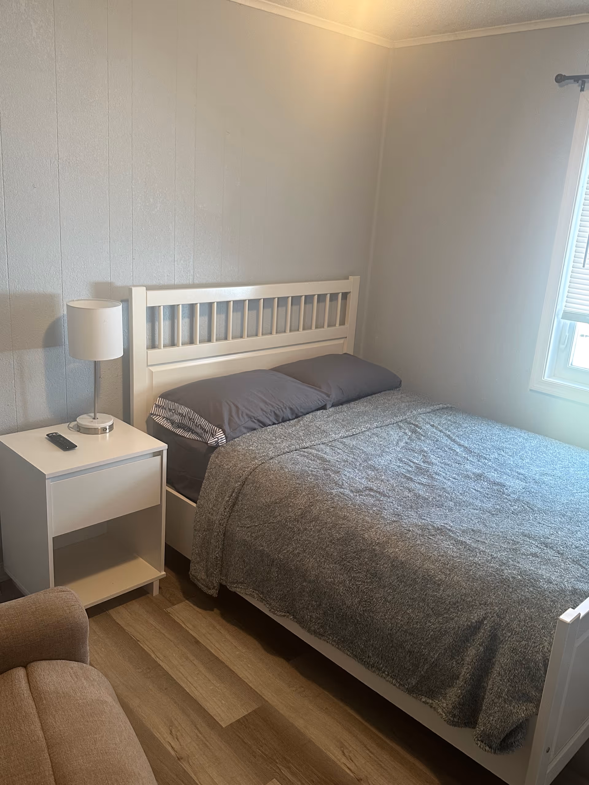 White bed with gray bedding and nightstand with lamp in a small, bright bedroom.