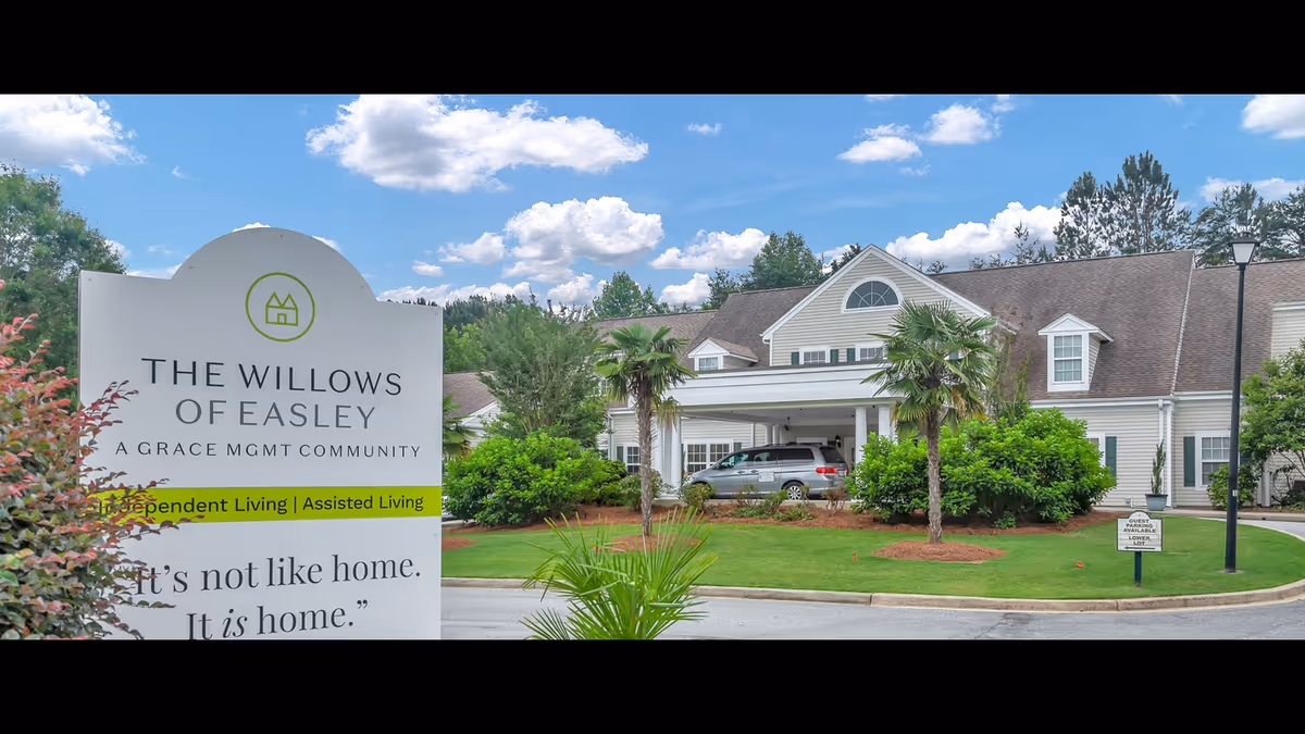 Exterior view of The Willows of Easley senior living facility showing a large white sign with the facility name and services offered, surrounded by greenery and a driveway with a parked vehicle under a covered entrance. The sky is blue with scattered clouds.
