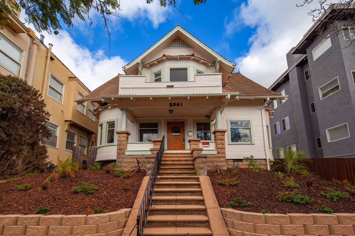 Front view of a white residential house with stairs, landscaping, and neighboring buildings.