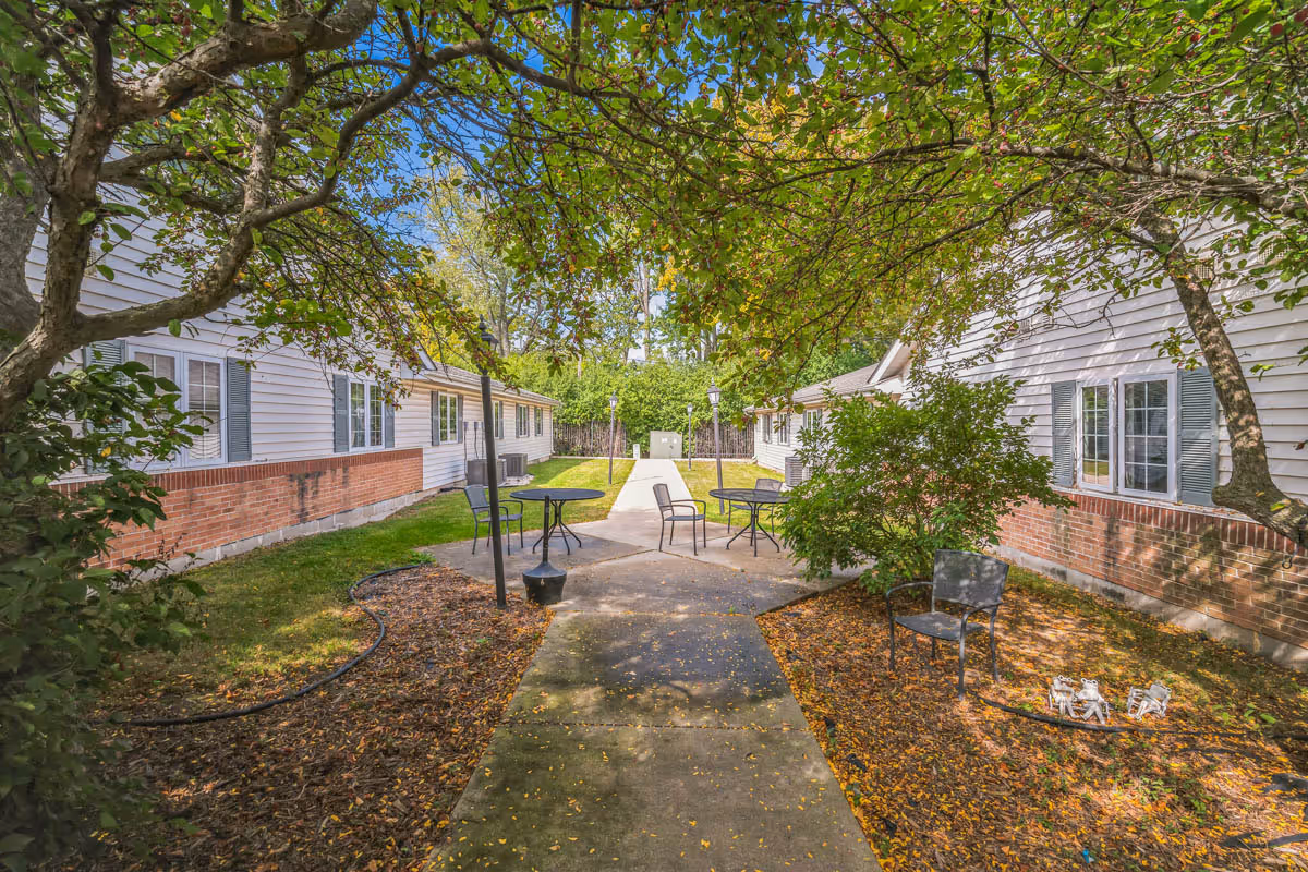 Outdoor courtyard area between two single-story buildings with white siding and brick accents. The courtyard has a concrete walkway, several metal tables and chairs, green grass, trees with green leaves, and some fallen leaves on the ground. The sky is clear and blue.