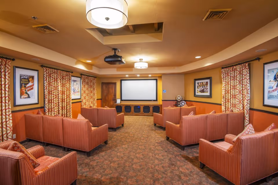 A cozy movie theater room with multiple rows of red upholstered chairs facing a large blank projection screen. The walls are decorated with framed vintage movie posters and patterned curtains. The ceiling has recessed lighting and two hanging light fixtures.