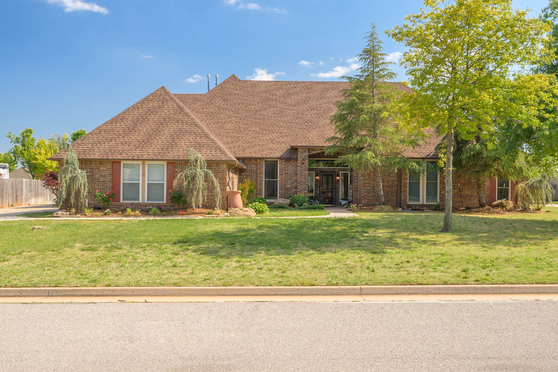 Front exterior view of a single-story brick building with a brown shingled roof, surrounded by green grass, trees, and shrubs under a clear blue sky.