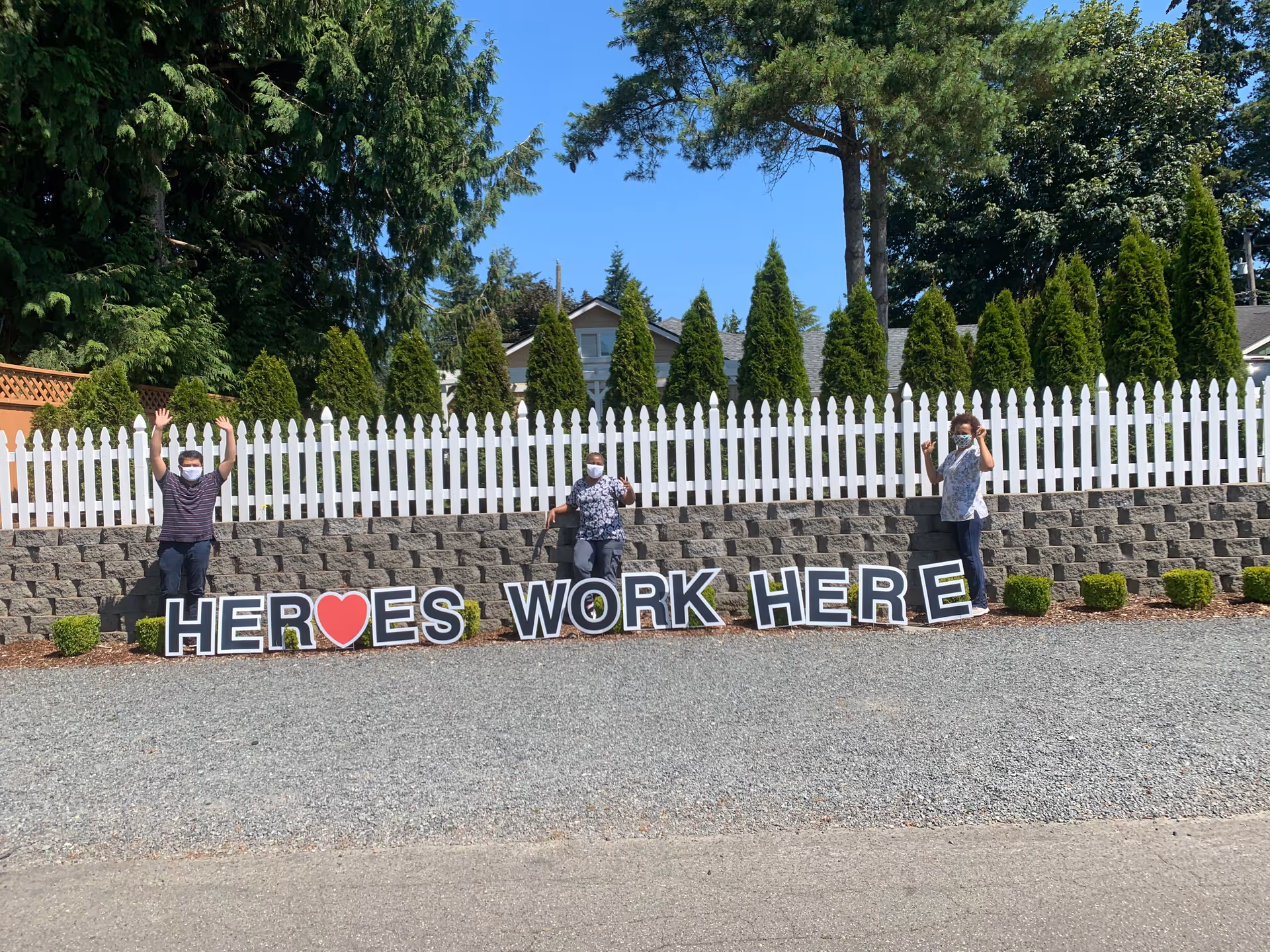Three masked people stand by a Heroes Work Here sign before a white picket fence.