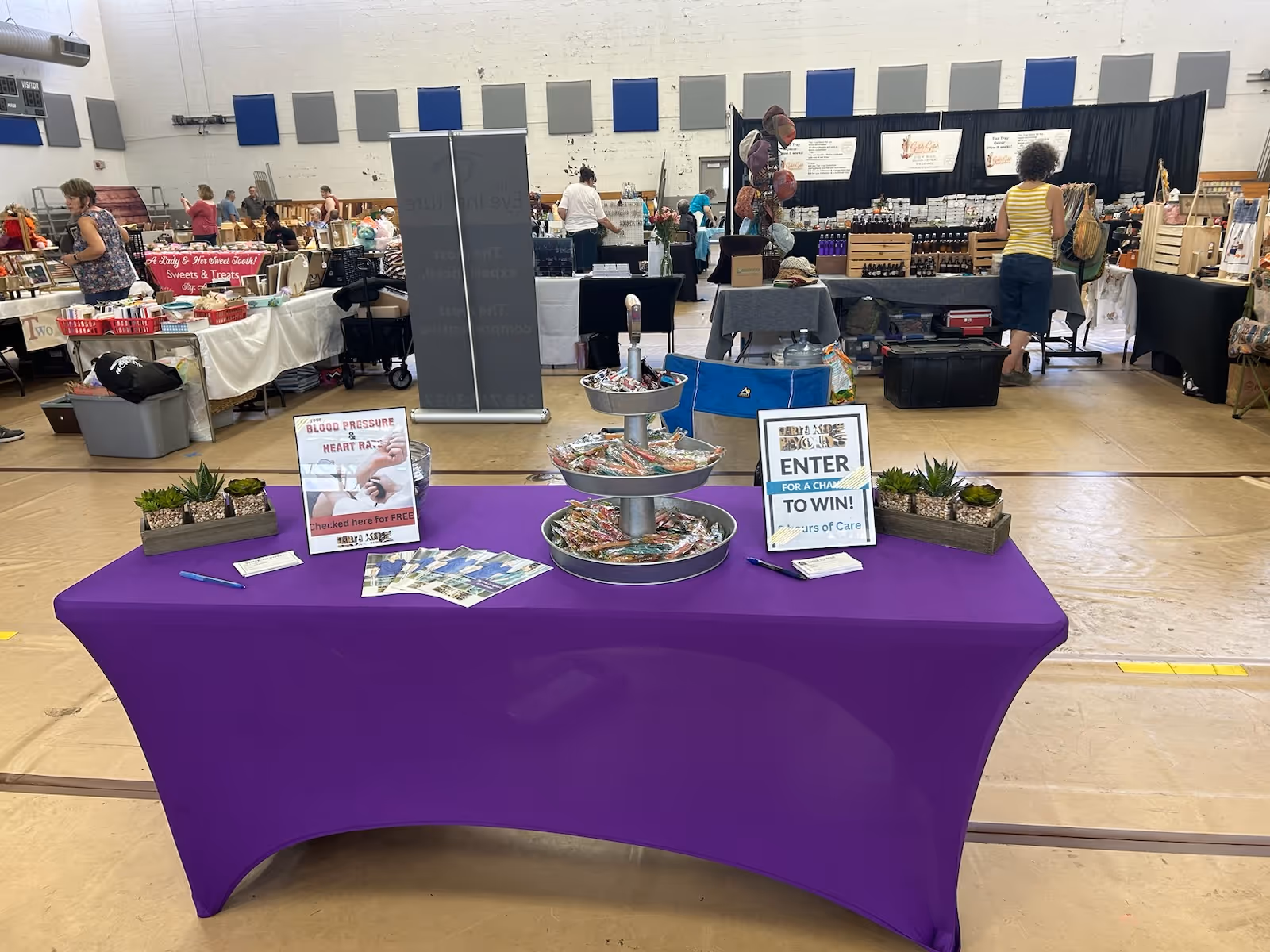 A purple table in a gym hosting a community fair, with vendor booths in background.
