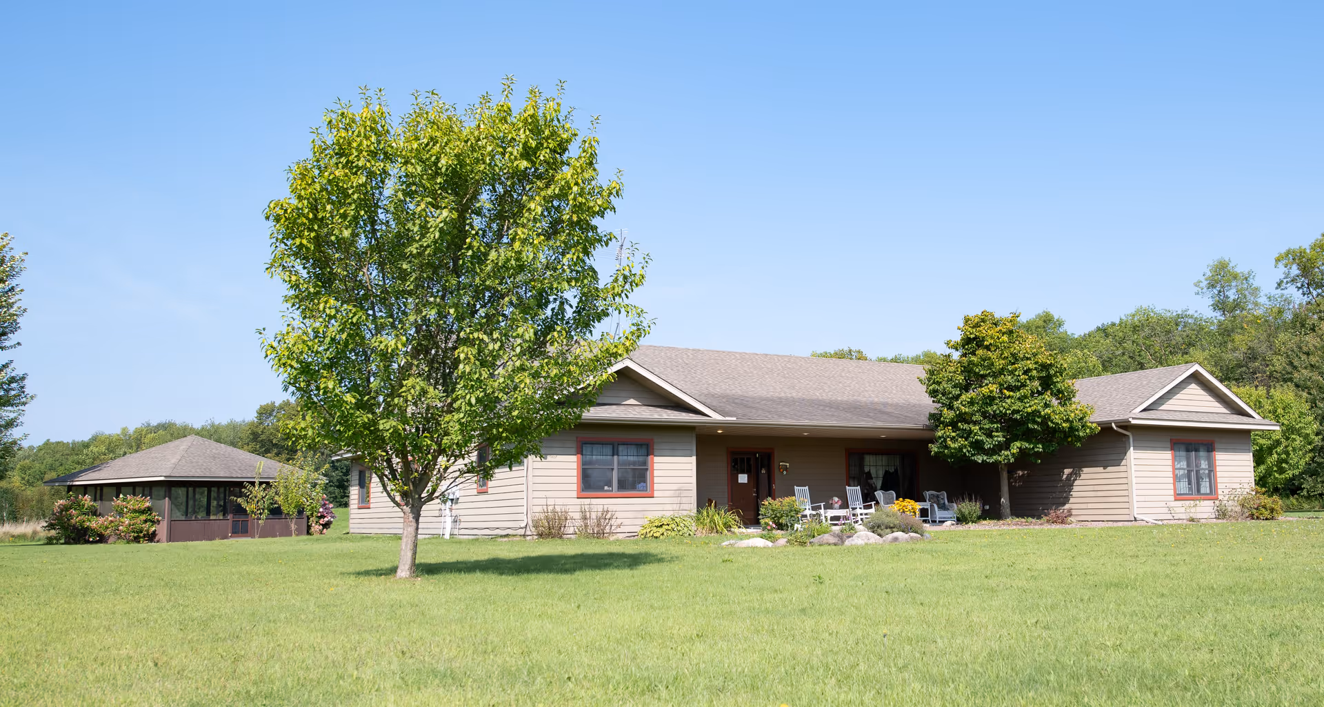 Single-story senior living facility building with a large lawn, trees, and a covered front porch with chairs under a clear blue sky.