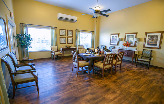 A spacious dining room with wooden flooring, a round dining table set with six chairs, and place settings. The room has light yellow walls, two large windows with curtains, framed artwork on the walls, a ceiling fan with a light fixture, and additional chairs and a side table along the walls.