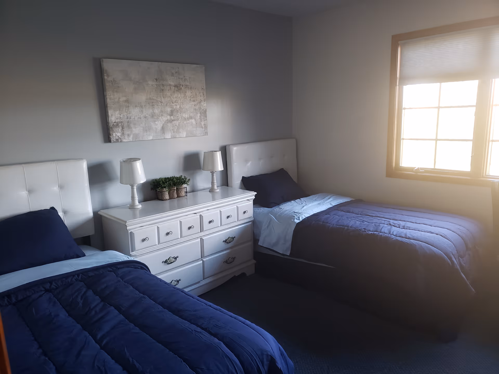 Two twin beds with blue quilts flank a white dresser in a bright, softly lit bedroom.