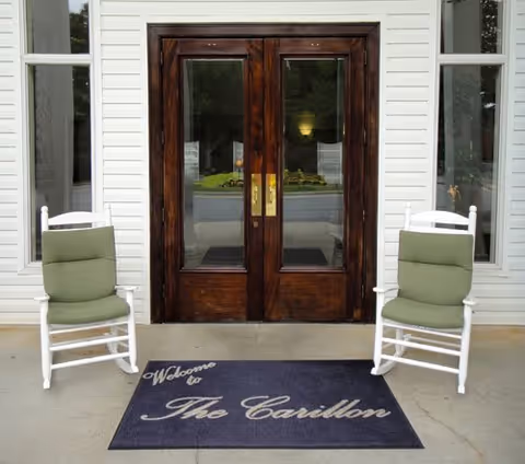 Wooden double doors with white siding, two green chairs, and a welcome mat.