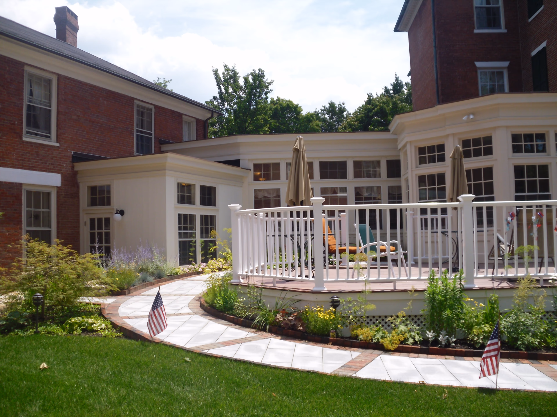 Outdoor patio area with white railing and several chairs and umbrellas, surrounded by a garden with green plants and small American flags, adjacent to a brick building with many windows.