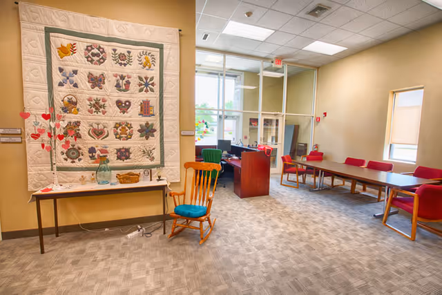 Quilt hangs on wall beside rocking chair and long table with red chairs in bright interior