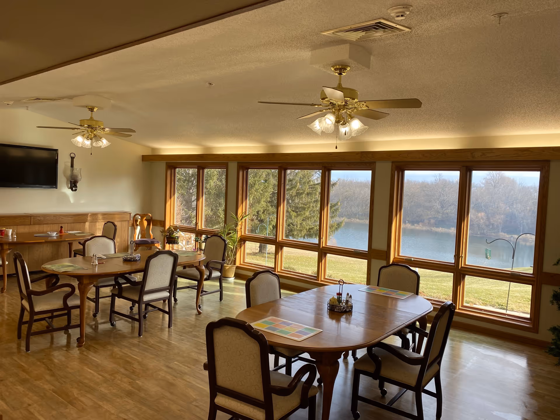 A dining room with wooden tables and cushioned chairs arranged neatly. Large windows provide a view of a lake and trees outside. The room has ceiling fans with lights and a wall-mounted television.