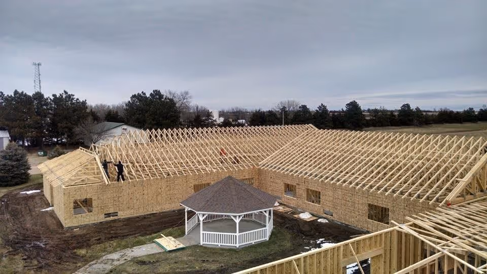 A large building under construction with wooden framing and roof trusses visible. The structure is in an L-shape surrounding a small white gazebo in the center. The surrounding area has some trees and open land under a cloudy sky.
