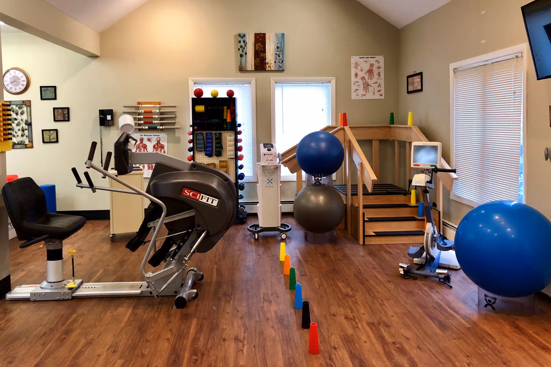 A physical therapy or exercise room with various fitness equipment including a SciFit exercise machine, large blue and gray exercise balls, a wooden ramp with stairs, colorful cones lined up on the wooden floor, and anatomical charts on the walls. The room has two windows with blinds and light green walls.