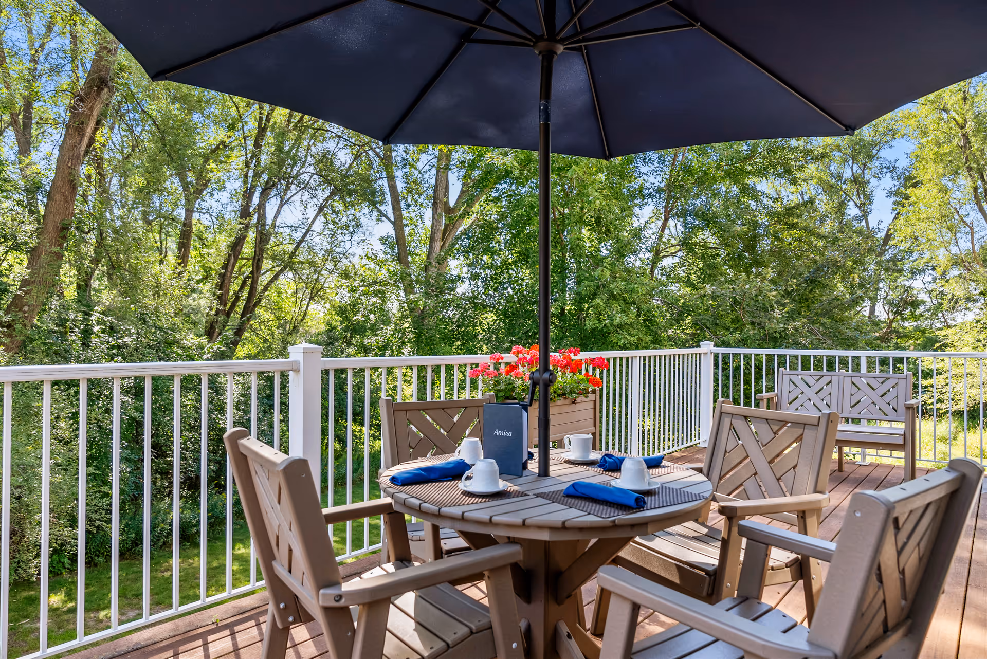 Outdoor deck with a round table, chairs, umbrella and potted flowers overlooking a wooded area.