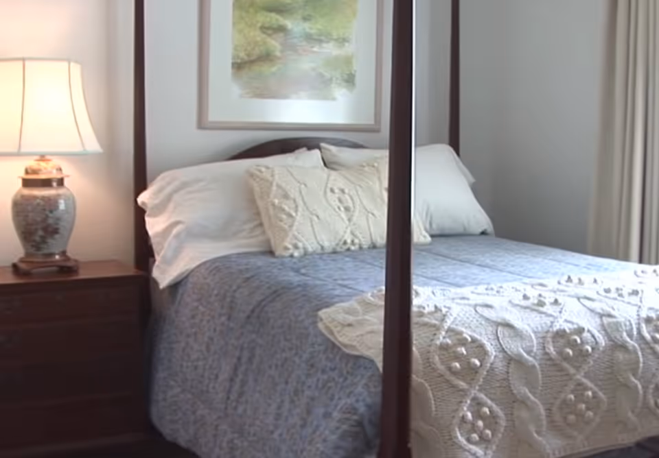 A cozy bedroom featuring a wooden four-poster bed with white pillows and a blue and cream knitted blanket. A wooden nightstand with a decorative lamp is positioned to the left of the bed, and a framed landscape painting hangs on the wall above the headboard.