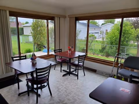 A small dining area with three dark wooden tables and several chairs. Each table has a small centerpiece, and large windows provide a view of a green yard with trees and neighboring houses. A walker is positioned near one of the tables.