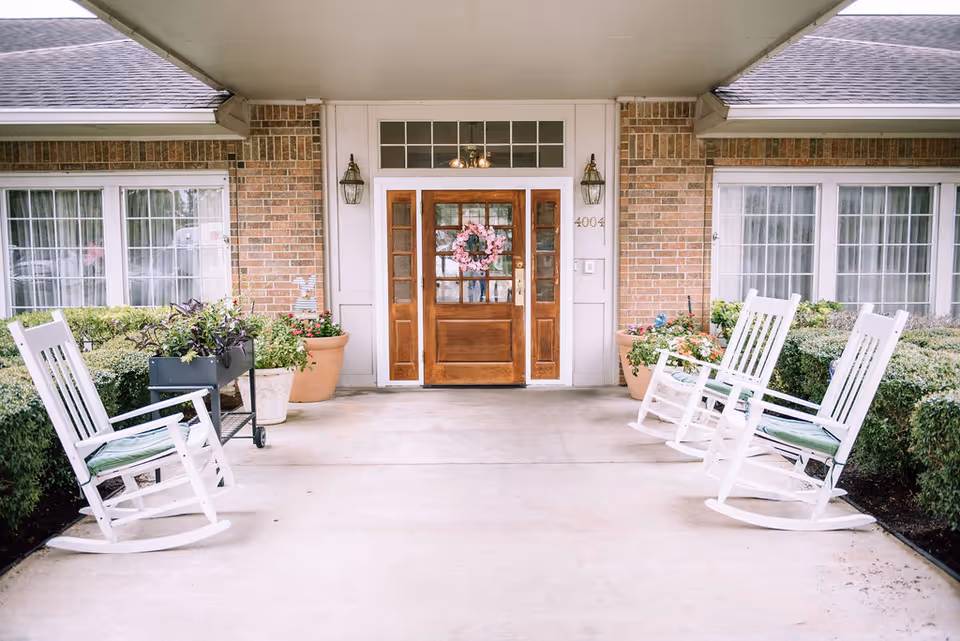 Covered entrance of a brick building with a wooden door decorated with a pink floral wreath. There are two large windows with white curtains on either side of the door. Four white rocking chairs with cushions are placed on the concrete porch, two on each side. Potted plants and greenery line the sides of the porch.