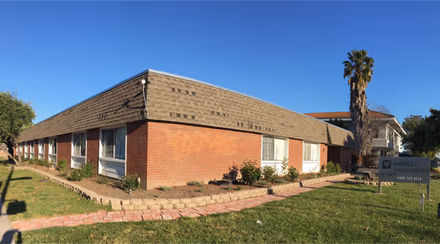 Single-story brick senior living building with a low-pitched roof, front lawn and a sign by the entrance under a clear blue sky.