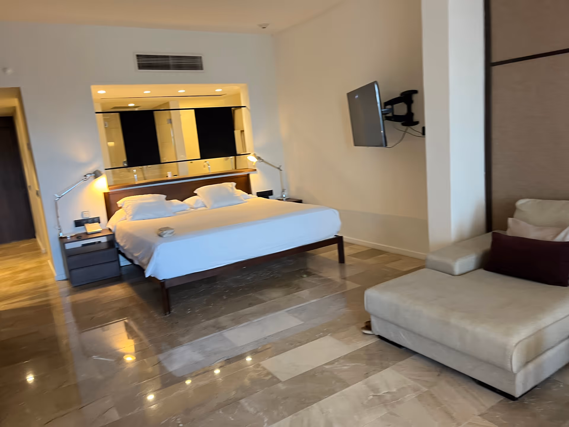 A modern hotel-style bedroom with a king bed, seating area, and wall-mounted TV.