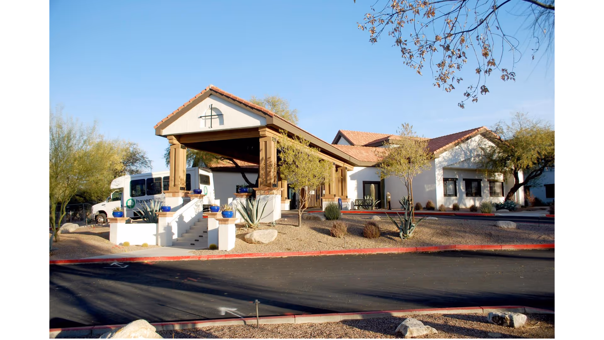 Exterior view of Haven Health Tucson facility showing a building with a covered entrance supported by wooden columns, desert landscaping with small trees and shrubs, and a parked white van with the Haven Health logo.