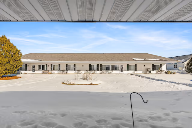Snow-covered parking area in front of a single-story beige senior living building under a blue sky