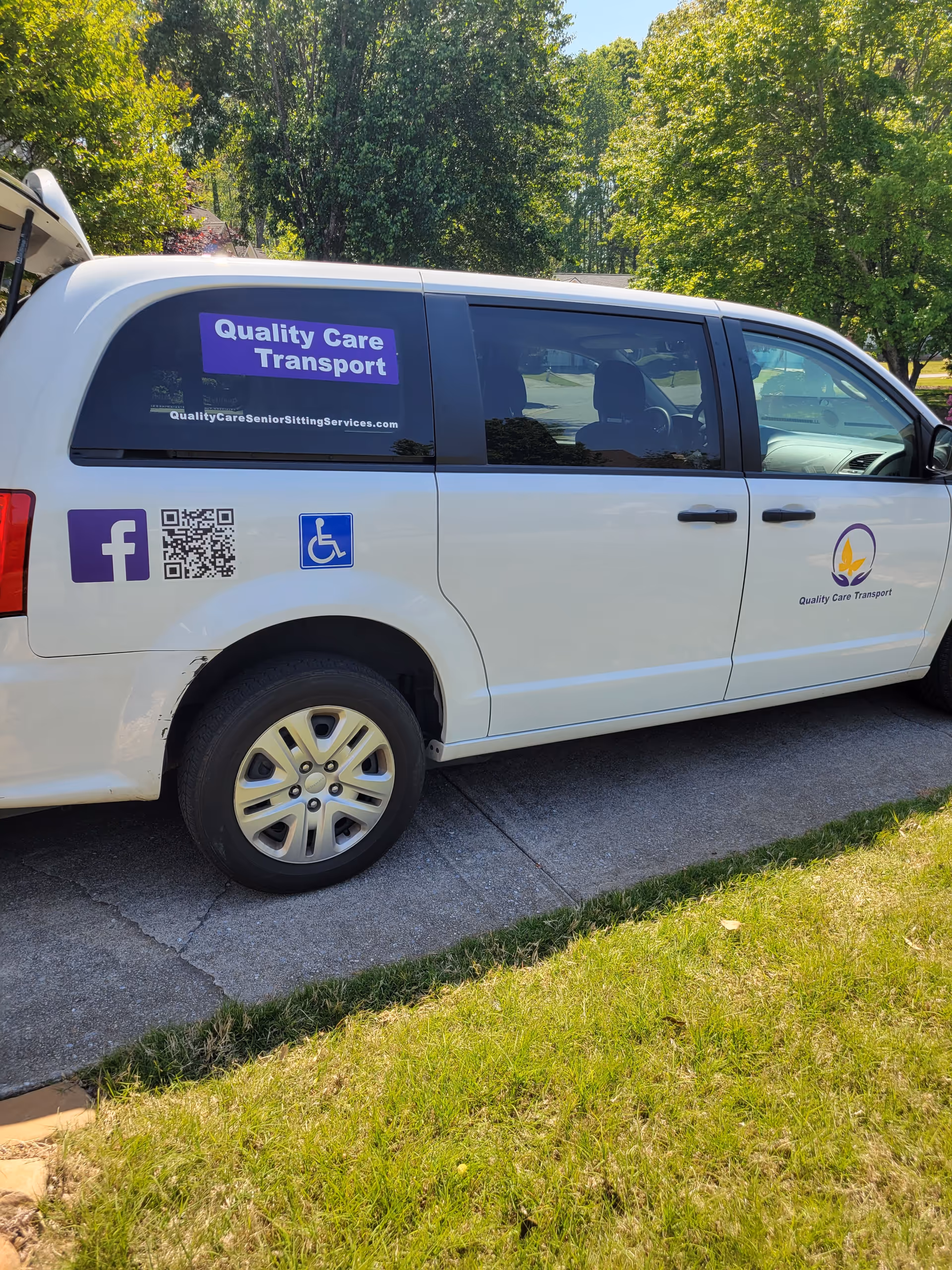 White transport van with Quality Care Transport signage parked outdoors near trees.