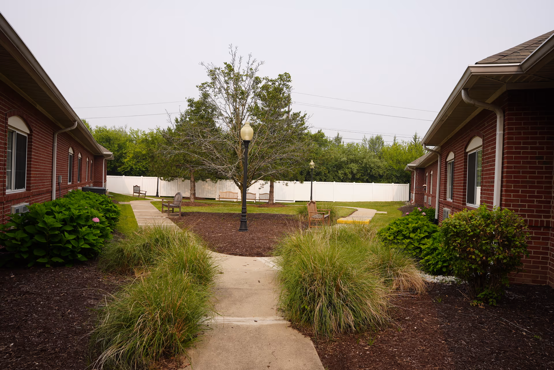 Outdoor courtyard area between two single-story brick buildings with a concrete walkway, green bushes, ornamental grasses, benches, a tree, and lamp posts, enclosed by a white fence with trees in the background.