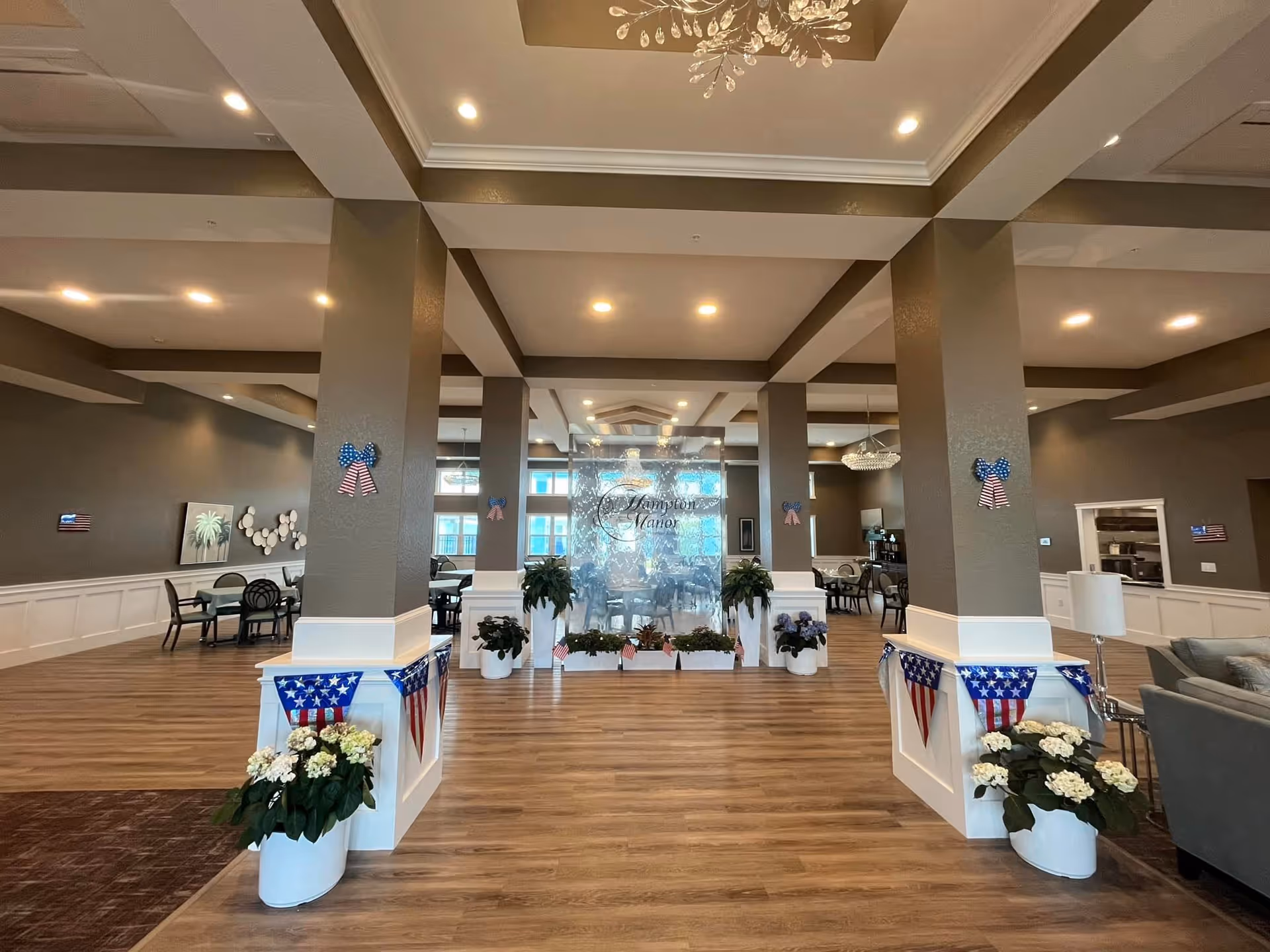 Spacious and well-lit interior of Hampton Manor Winchester Premier Assisted Living Facility featuring a large open area with wooden flooring, multiple seating arrangements with tables and chairs, decorative plants, patriotic American flag-themed decorations, and a central glass panel with the facility's name. The ceiling has recessed lighting and a decorative chandelier.