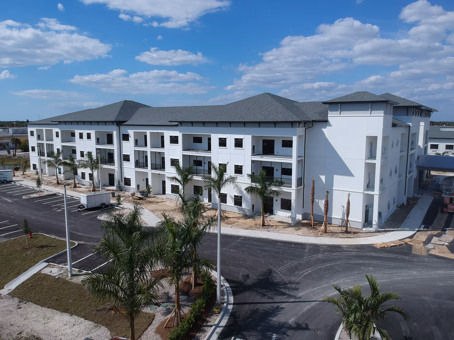 Exterior view of a three-story white building with a gray roof, surrounded by newly planted palm trees and a paved parking lot under a partly cloudy blue sky.