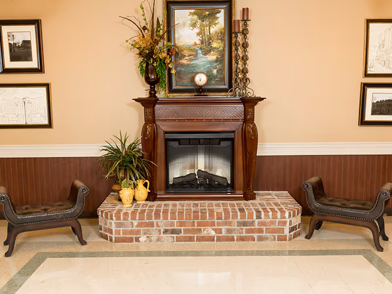 A cozy indoor setting featuring a brick fireplace with a wooden mantel. On the mantel, there is a framed painting of a river landscape, a small clock, a tall candle holder with a candle, and a vase with dried flowers. On either side of the fireplace, there are two dark brown leather benches with nailhead trim. The walls are decorated with framed black and white sketches, and the lower half of the wall has wood paneling.