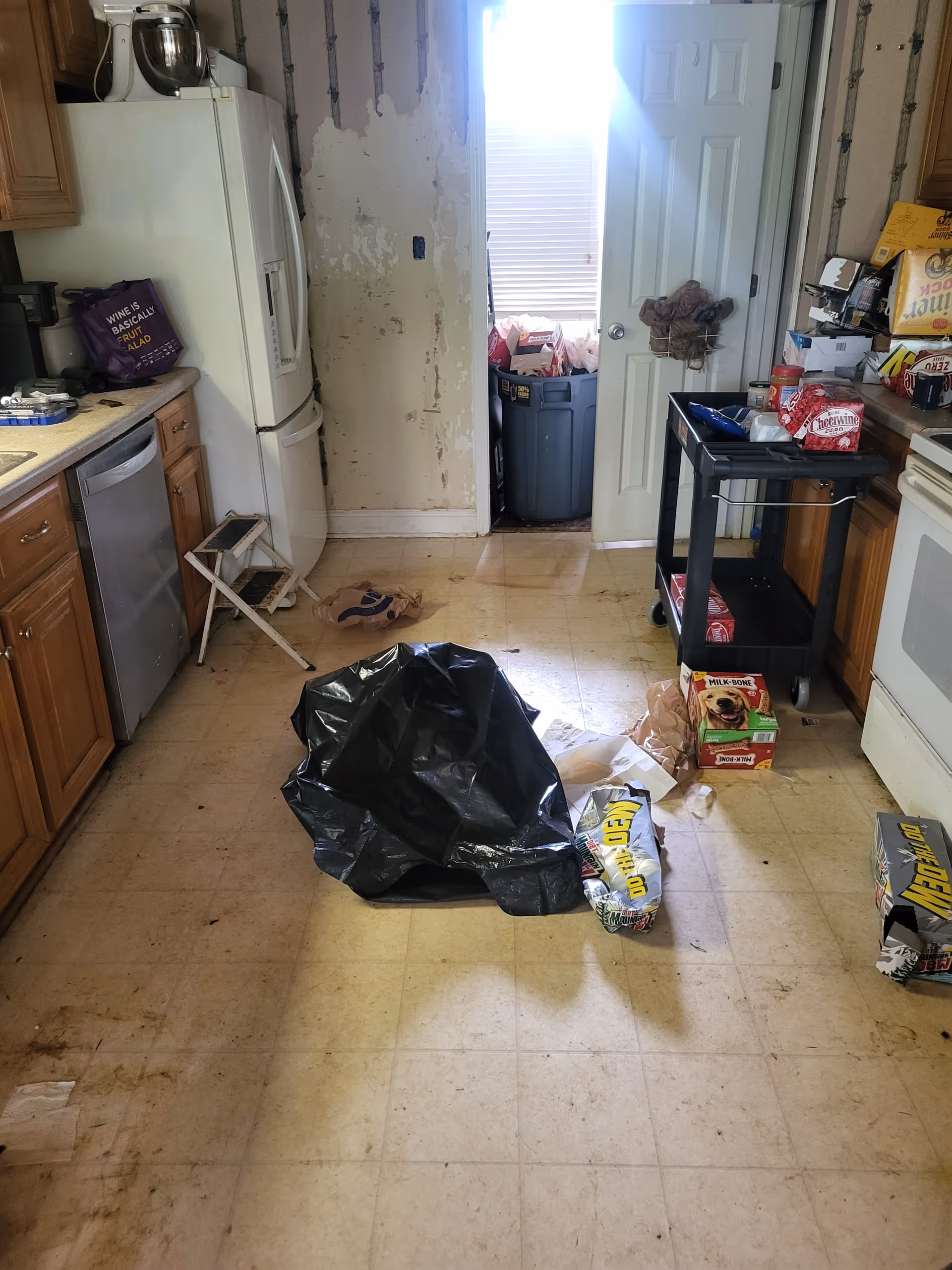 Messy kitchen with a black trash bag on the floor and a damaged wall.