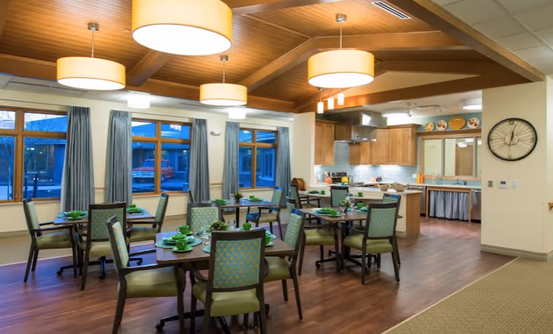 Bright communal dining room with multiple set tables and chairs, pendant lights, large windows, and an open kitchen area.