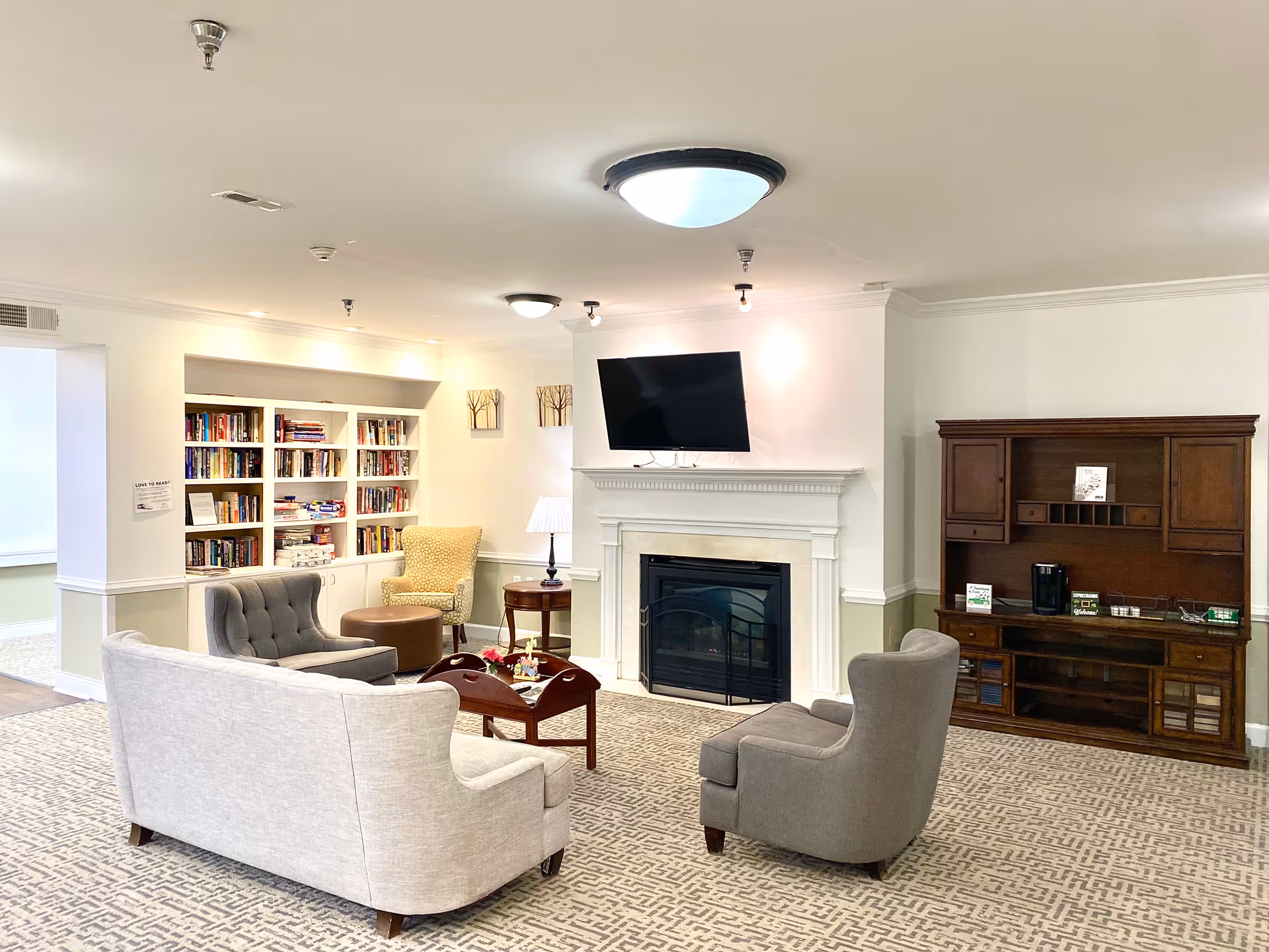 Comfortable senior living room with sofas and armchairs centered around a fireplace and wall-mounted TV, with bookshelves and cabinets along the walls.