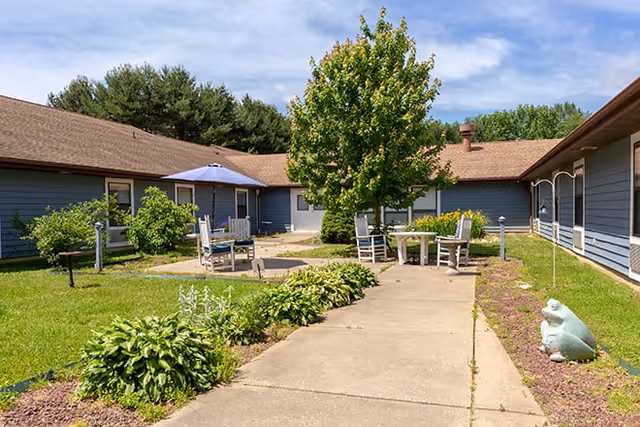 Outdoor courtyard area at Brookdale Dover with a concrete walkway, green grass, plants, a tree in the center, several chairs and tables, and a blue umbrella under a clear sky.