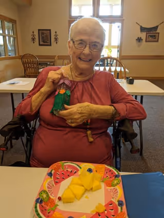 An elderly woman sitting in a wheelchair at a table in a well-lit room, smiling and holding up a small colorful craft. On the table in front of her is a plate with pieces of cut pineapple. The room has wooden chairs and tables, with windows and framed decorations on the walls.