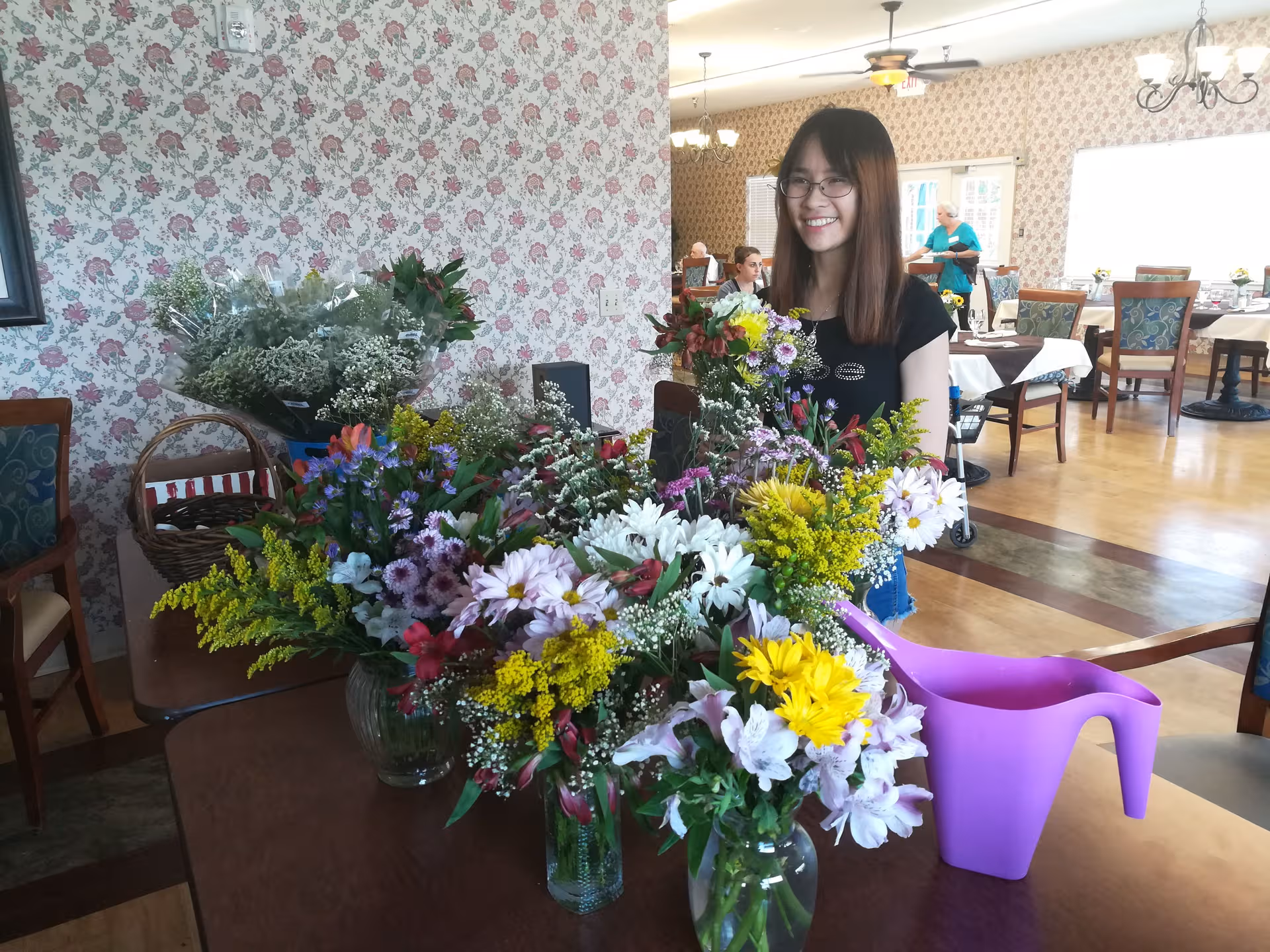 A smiling woman stands behind a large bouquet on a dining room table.