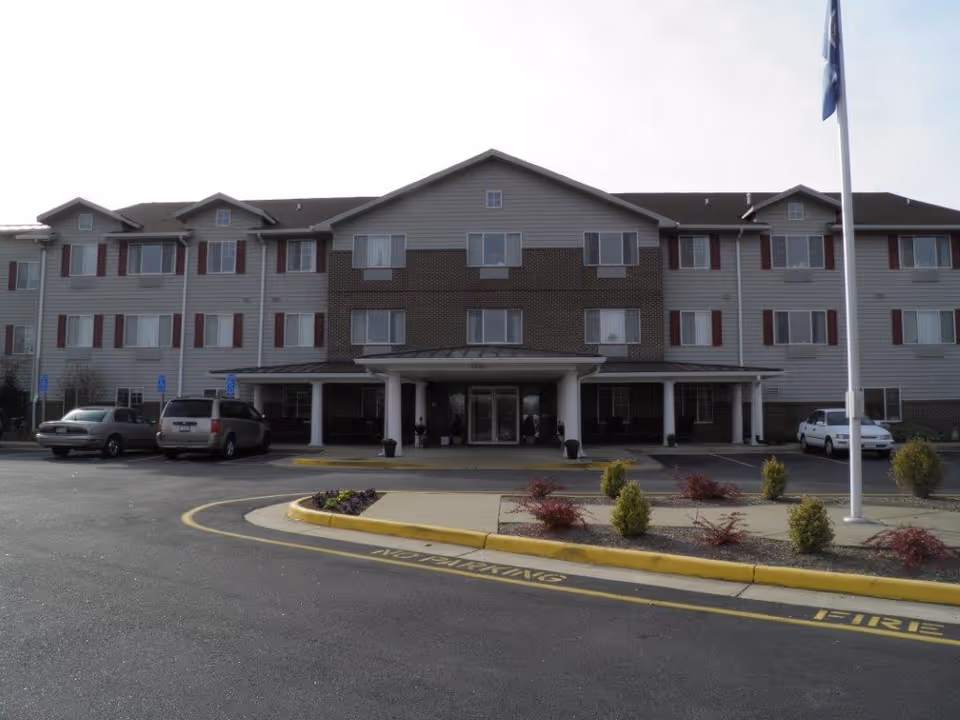 Three-story senior living building front with a covered entrance, drive-up roundabout, parked cars, flagpole and landscaped islands.
