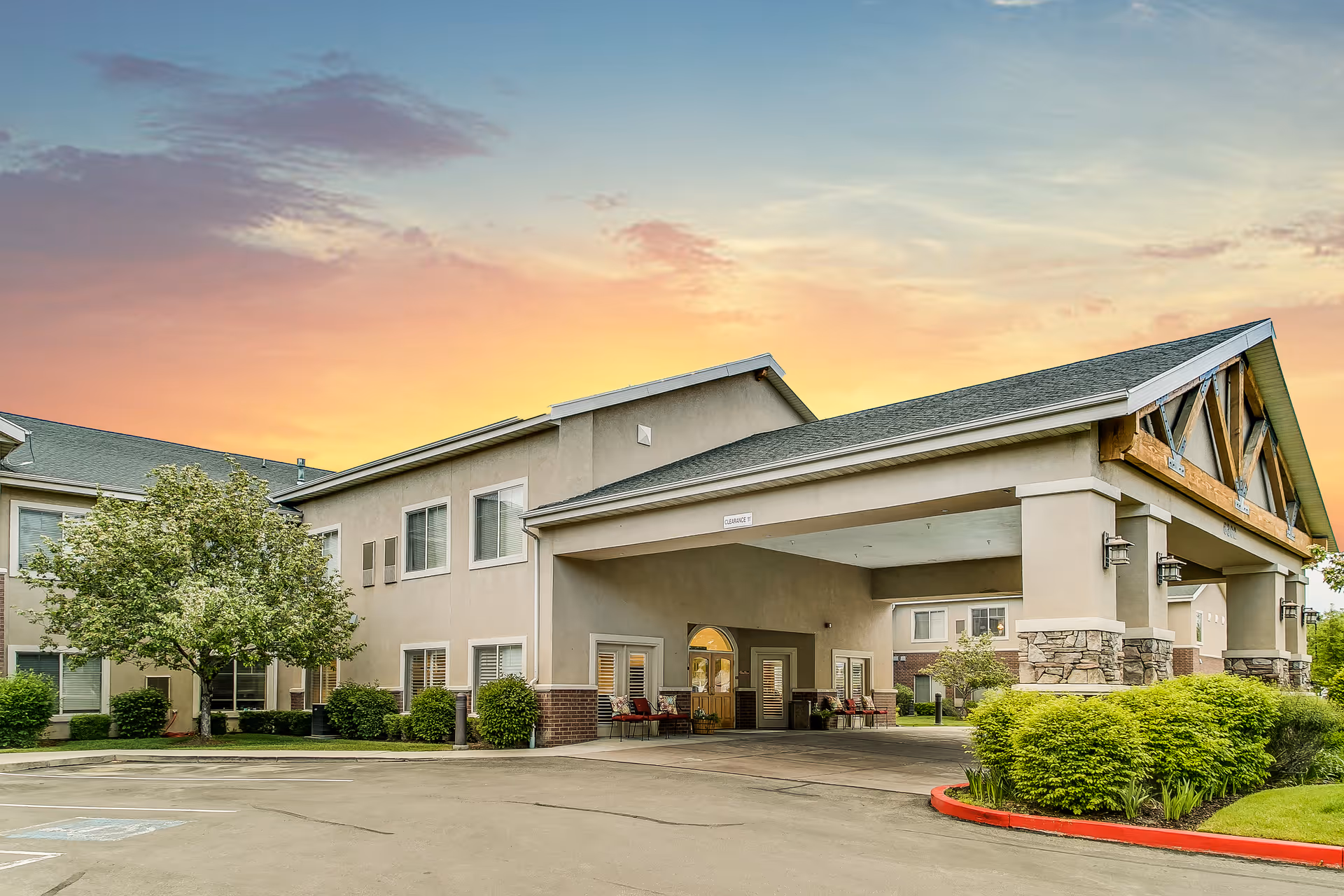 Front entrance of a senior living building with a covered porte-cochere, landscaped shrubs, and a colorful sunset sky.