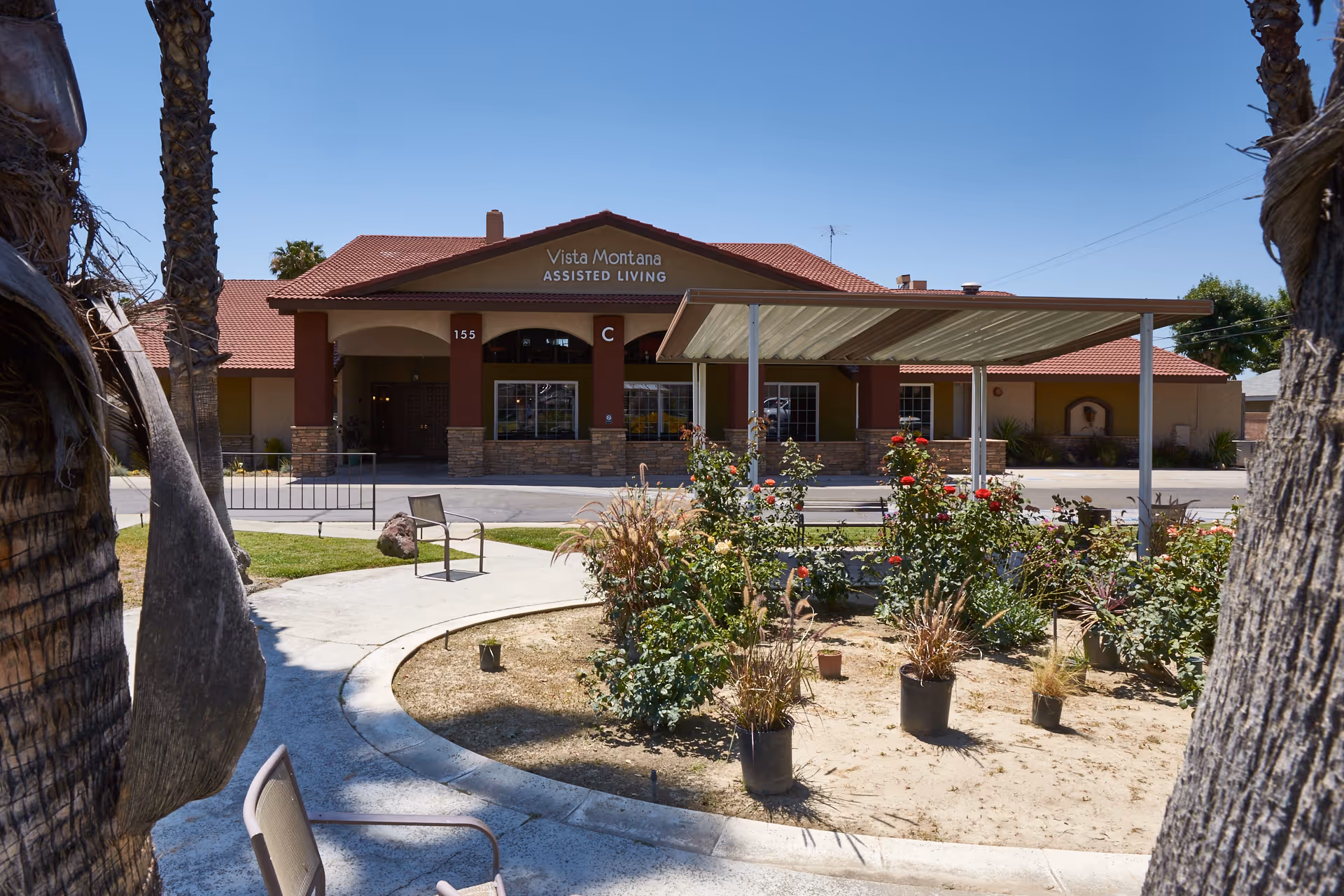 Exterior view of Vista Montana Assisted Living facility with a covered entrance, garden area with plants and flowers, and a clear blue sky.