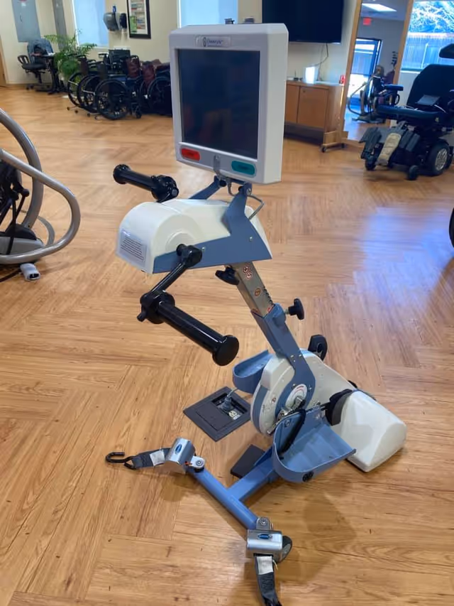 A motorized exercise machine with handles and foot pedals, designed for physical therapy or rehabilitation, placed on a wooden floor in a room with wheelchairs and other medical equipment in the background.
