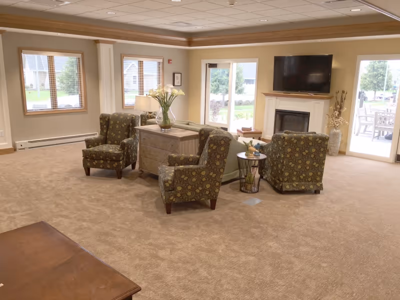 A spacious living room area with beige carpet and walls, featuring three patterned armchairs arranged around a small table and a wooden chest with a vase of flowers. There is a fireplace with a flat-screen TV mounted above it, large windows with blinds, and glass doors leading to an outdoor patio with seating.