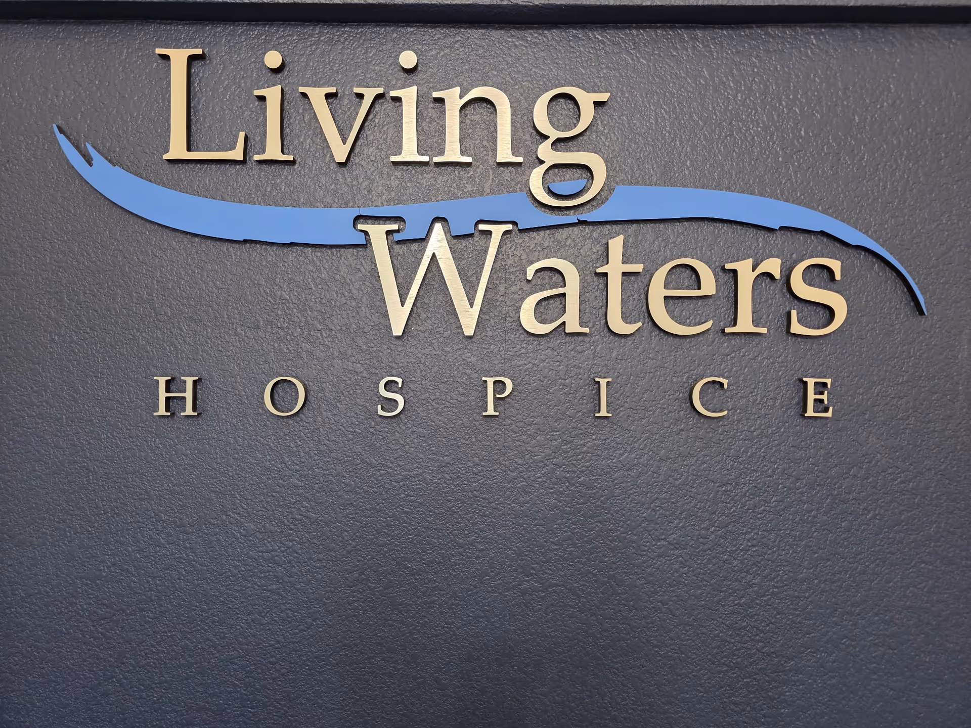 Close-up of a dark exterior wall sign reading Living Waters Hospice with a blue wave