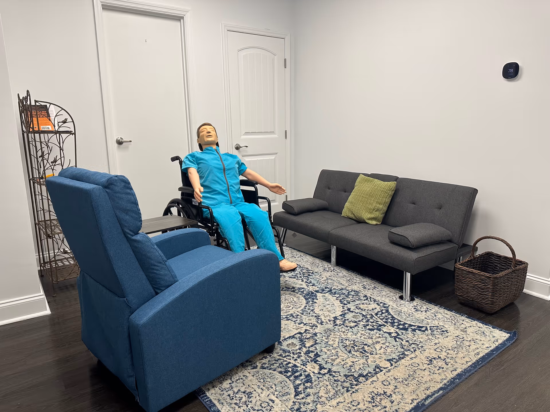 Mannequin in teal scrubs sits in a wheelchair beside a blue recliner and gray sofa.