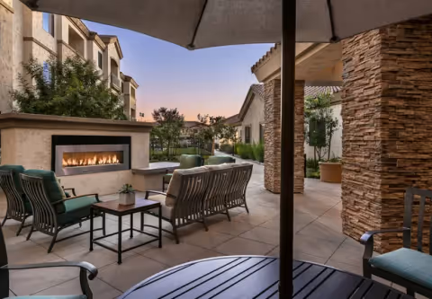 Outdoor patio area at a senior living facility with cushioned chairs and sofas arranged around a modern rectangular fireplace. Stone pillars and beige buildings surround the patio, with trees and plants in the background under a clear evening sky.